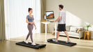 Two people using walking pads in a home setting with a computer monitor displaying a colorful screen