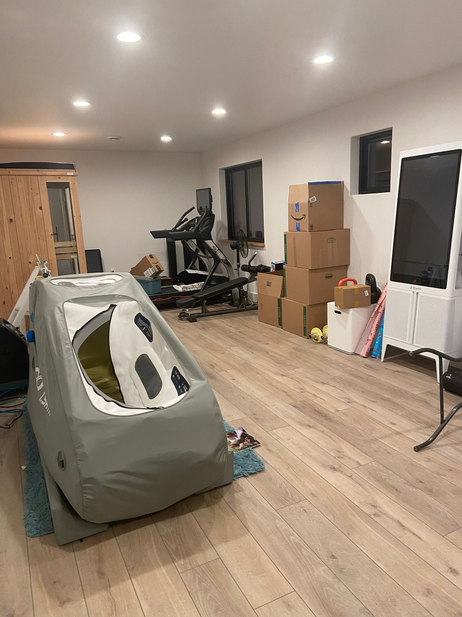 Home gym with treadmill, stacked boxes, hyperbaric chamber and a TV in a room with wooden flooring.
