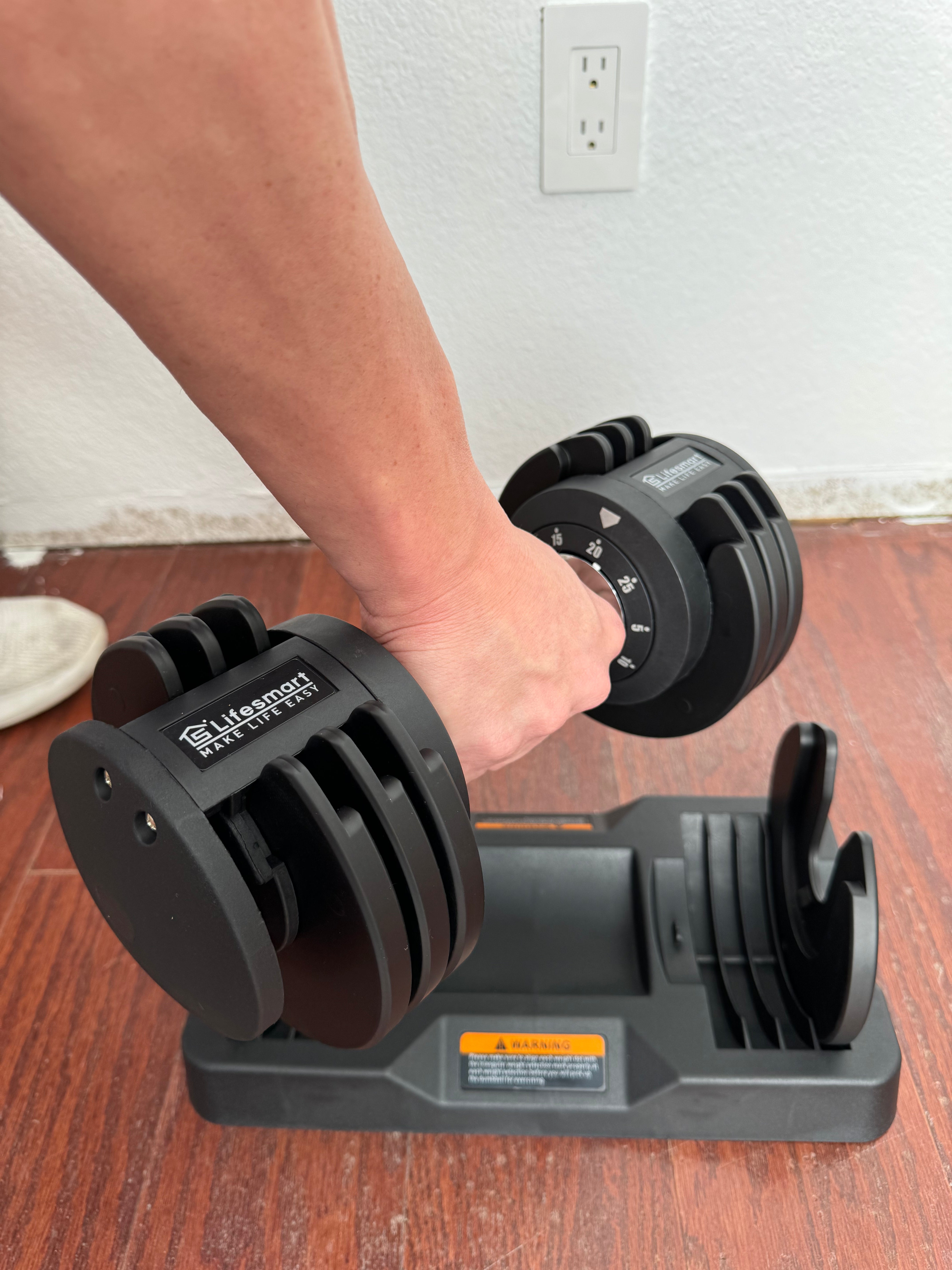 Person using a pair of adjustable dumbbells on a wooden floor.