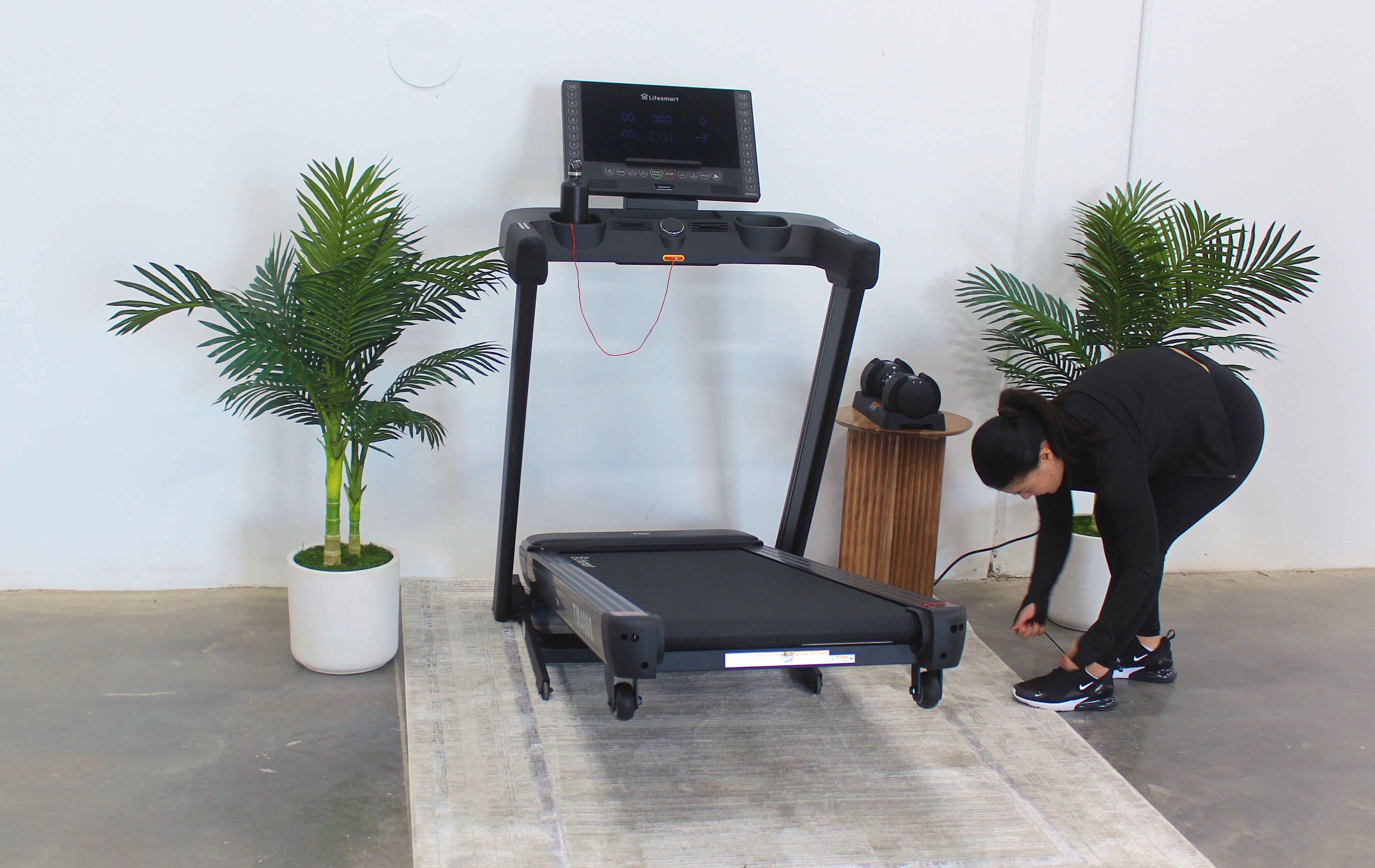 Person adjusting a treadmill in a room with plants and a rug.