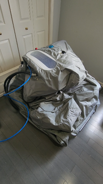 Sitting hyperbaric chamber deflated in a gray room on a wooden floor with accessory units attached